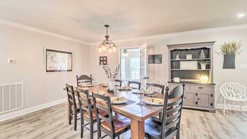 A dining area with a wooden table and chairs, a chandelier, and a kitchen area with cabinets and a shelf.
