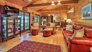 A cozy cabin interior with a wooden ceiling, a red leather couch, a wooden bookcase, and a dining area.