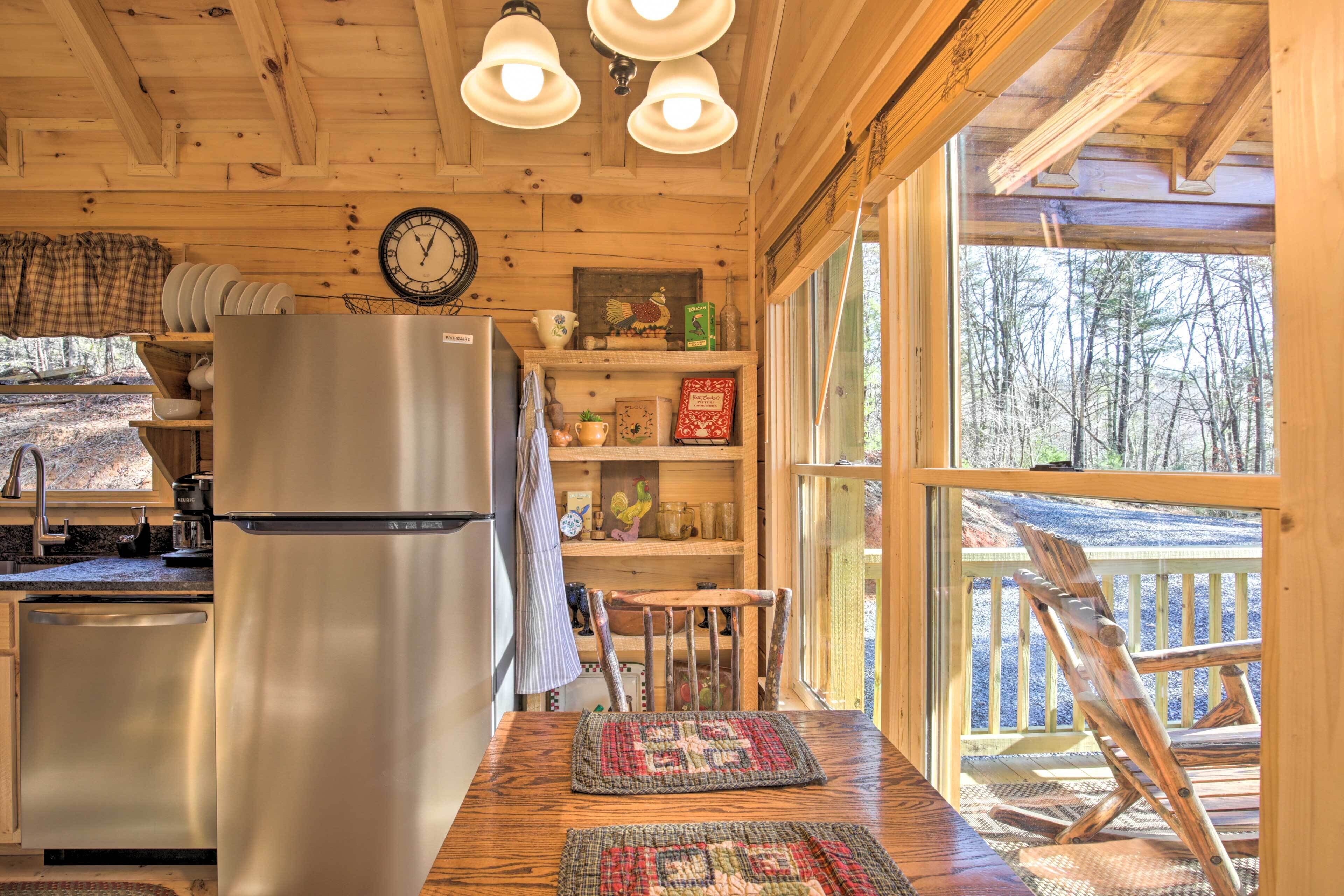 A cabin kitchen with a stainless steel refrigerator, wooden shelves, a clock, and a view of the forest through large windows.