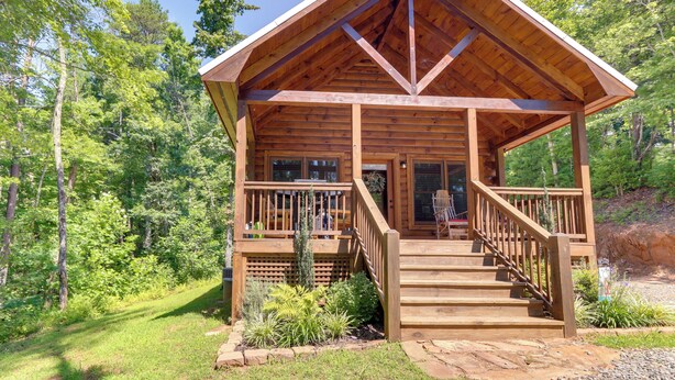 Une cabane en bois dotée d’un porche et d’un escalier, entourée de verdure.