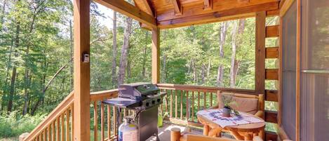 A wooden deck with a grill, a table, and chairs, surrounded by a forest.