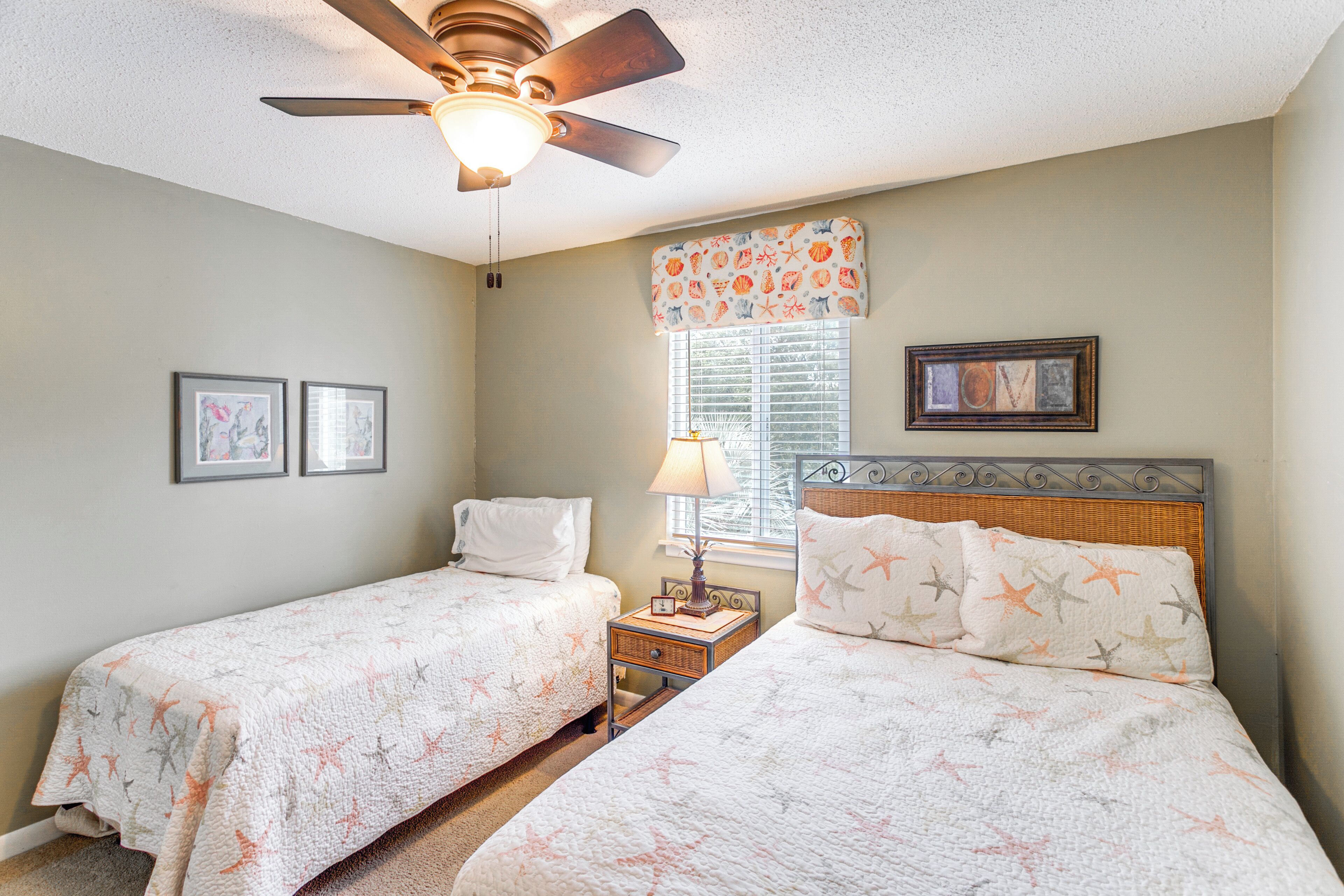 A bedroom with two beds, a ceiling fan, a nightstand with a lamp, and a window with blinds.
