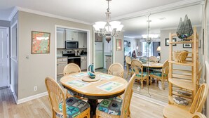 Uma sala de jantar com mesa redonda, cadeiras e lustre. Cozinha visível ao fundo, com armários e eletrodomésticos.