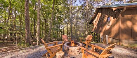 A rustic cabin with a fire pit surrounded by Adirondack chairs, set in a forested area.