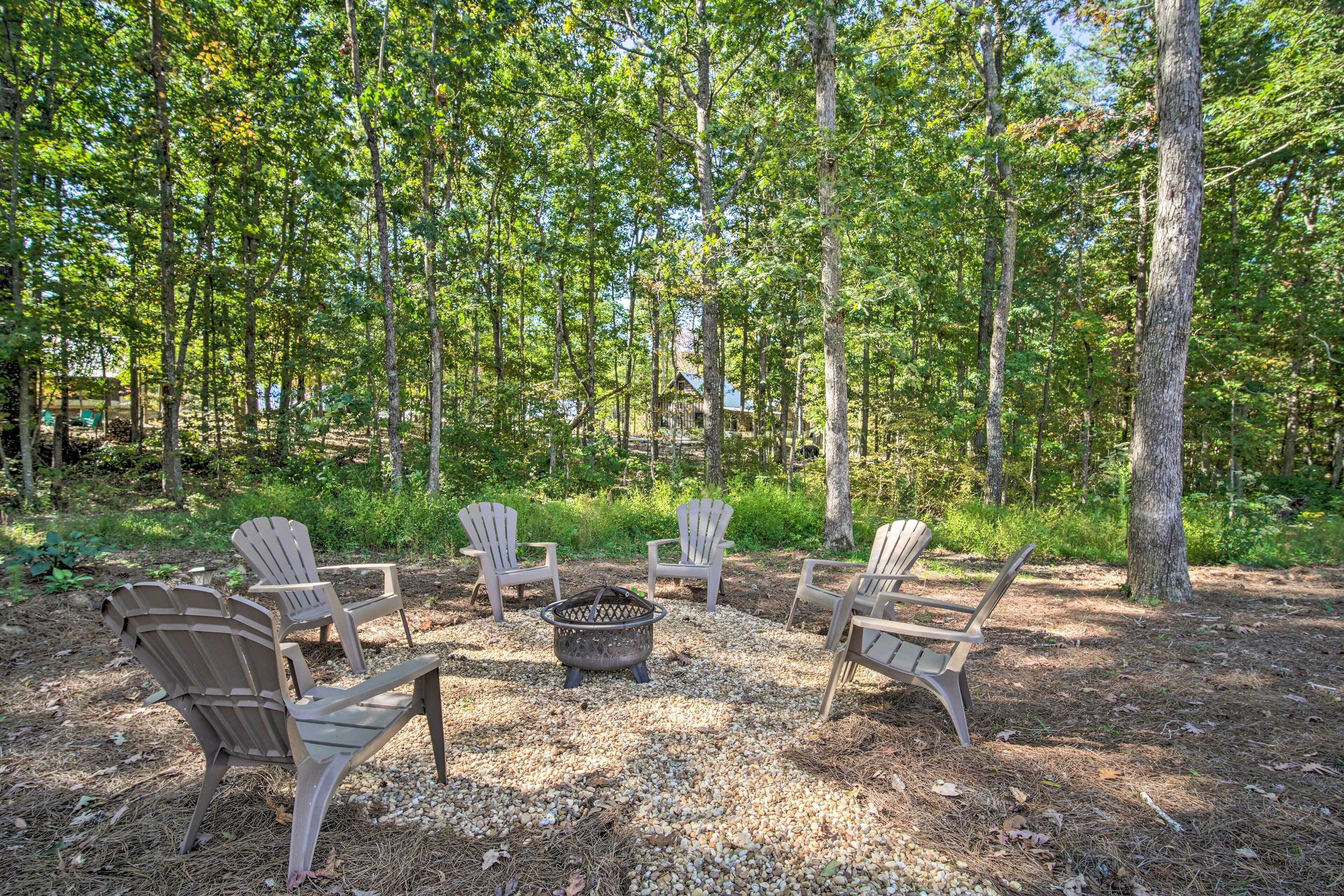 A forested area with Adirondack chairs arranged around a fire pit.
