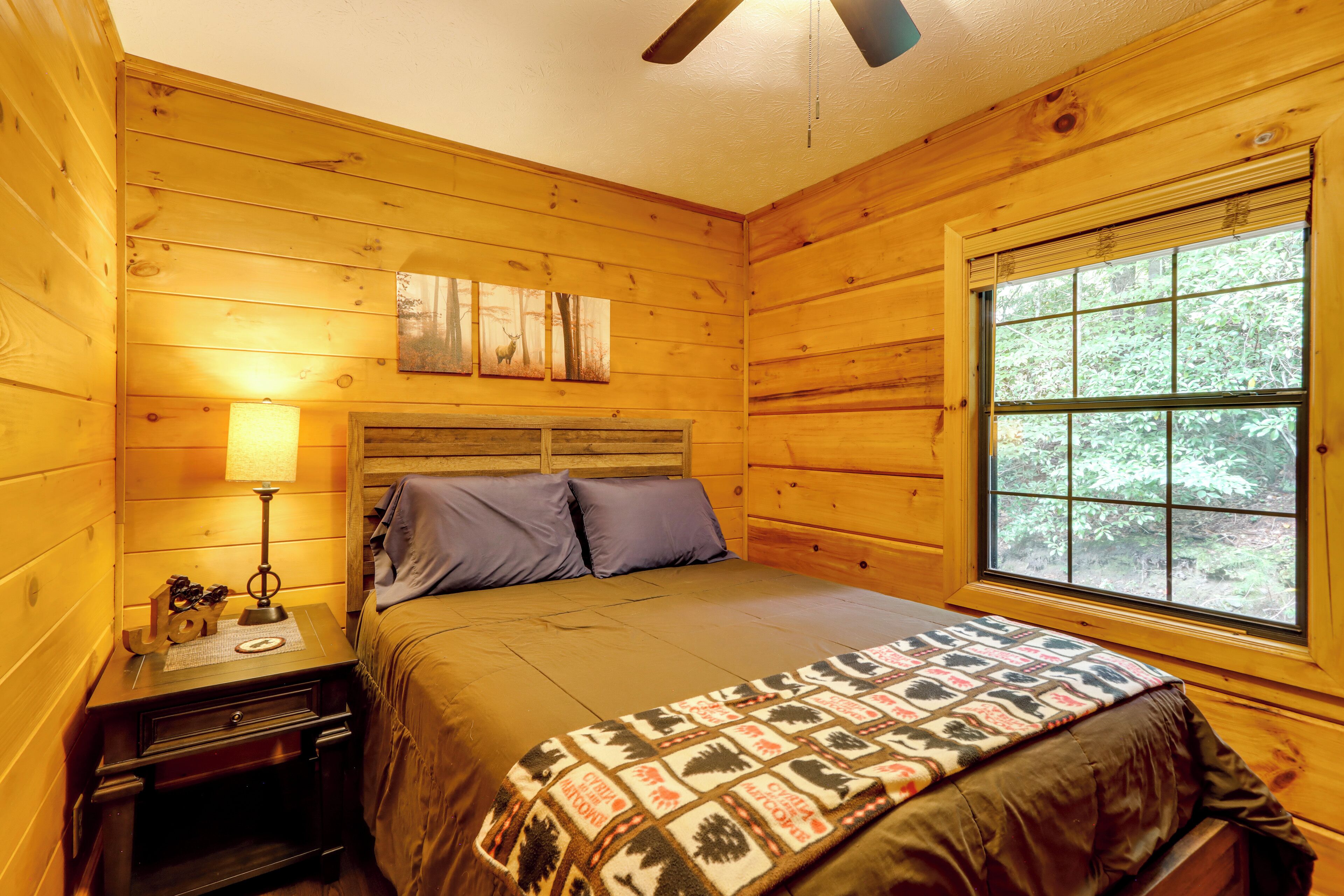 A wooden cabin bedroom with a bed, a nightstand, a lamp, and a window with a view of trees.