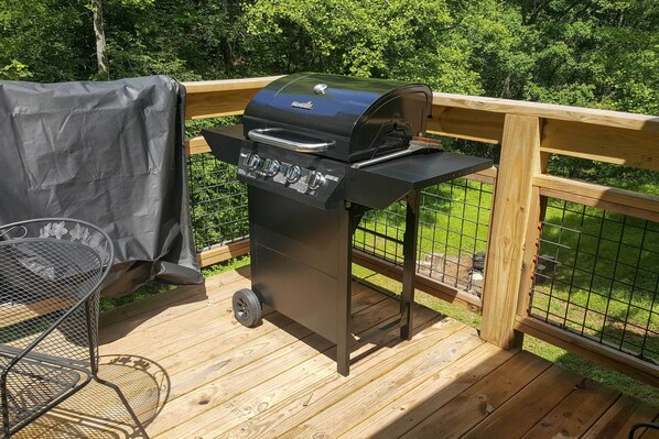 Una parrilla de barbacoa en una terraza de madera, con una silla metálica y una mesa cubierta cerca.