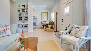 A living room with a sofa, coffee table, and decorative items. There is a dining area with a table and chairs. The room has white walls and a wooden floor. - Harbor Oaks Haven: Walk to Front Beach & Downtown! (Ocean Springs)
