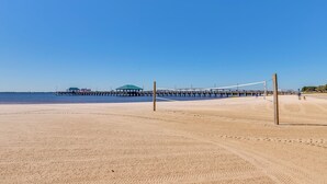 A beach volleyball court with a net, a wooden pier in the background, and a clear blue sky. - Harbor Oaks Haven: Walk to Front Beach & Downtown! (Ocean Springs)