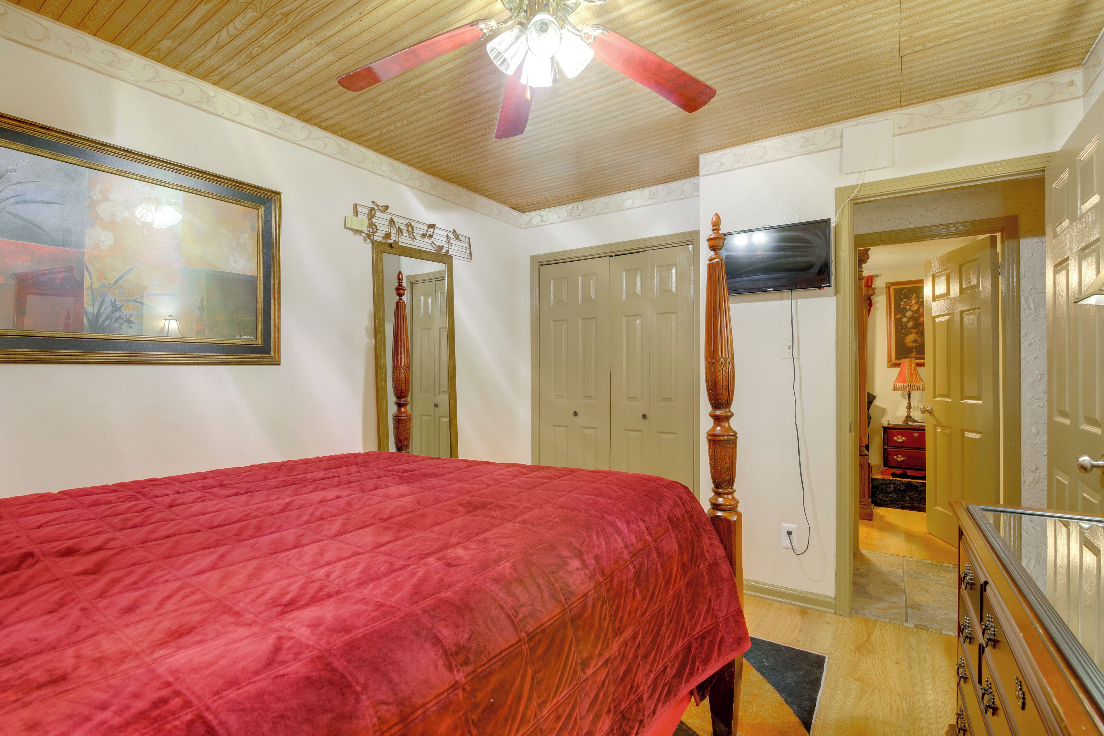 A bedroom with a red bedspread, a ceiling fan, a large mirror, and a door leading to another room.