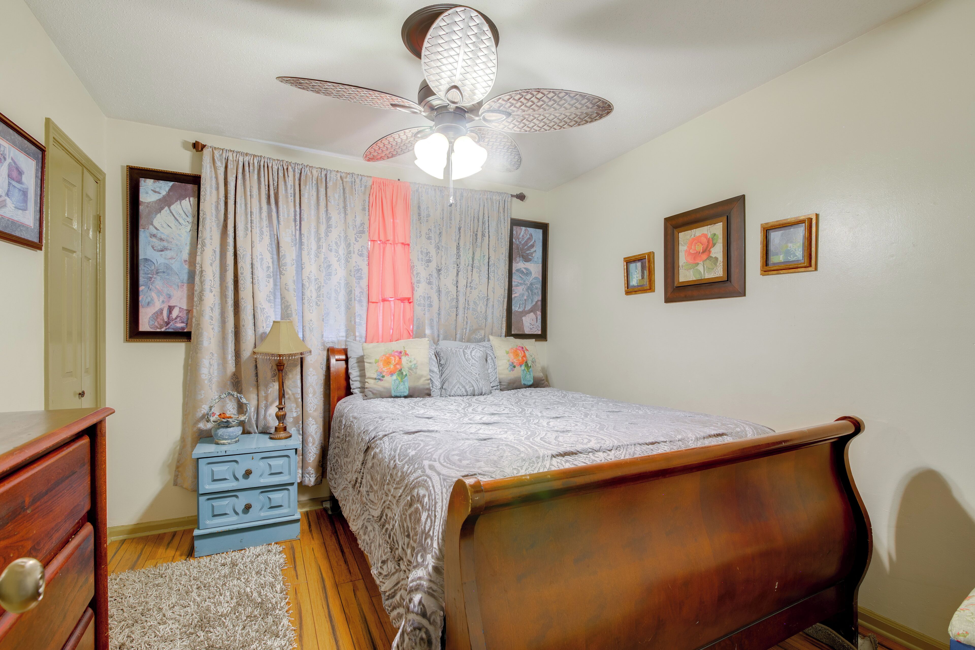 A bedroom with a wooden bed, a ceiling fan, a nightstand with a lamp, and a window with curtains.