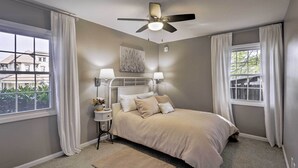 Une chambre à coucher avec un lit, deux lampes de chevet, un ventilateur de plafond et une fenêtre avec des rideaux.