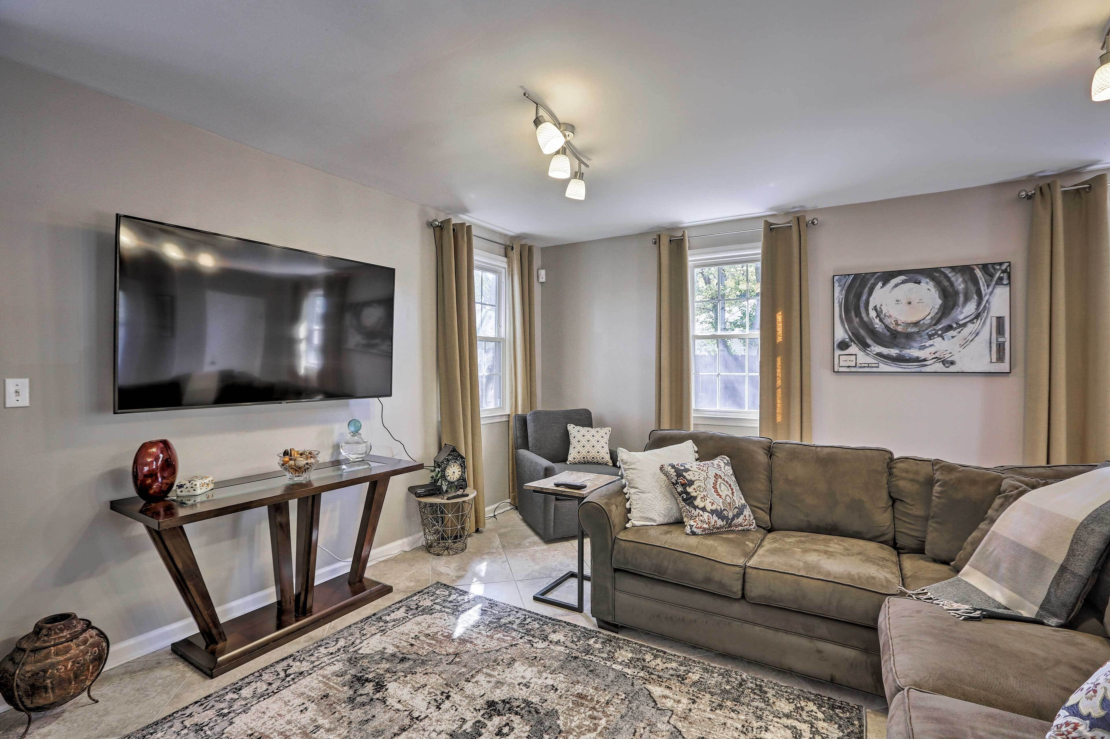 A living room with a large flat-screen TV, a dark wooden console table, a brown leather sofa, a patterned rug, and a painting on the wall.