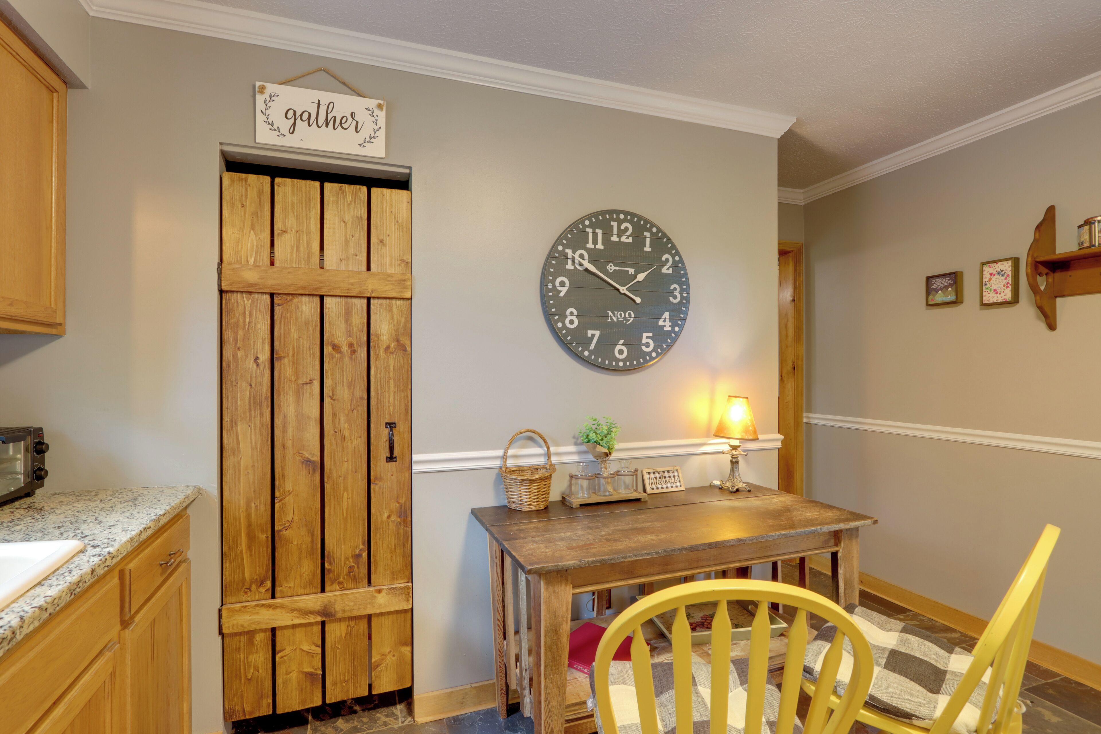 Una cocina con armarios de madera, un reloj, una mesa con una cesta y una silla.