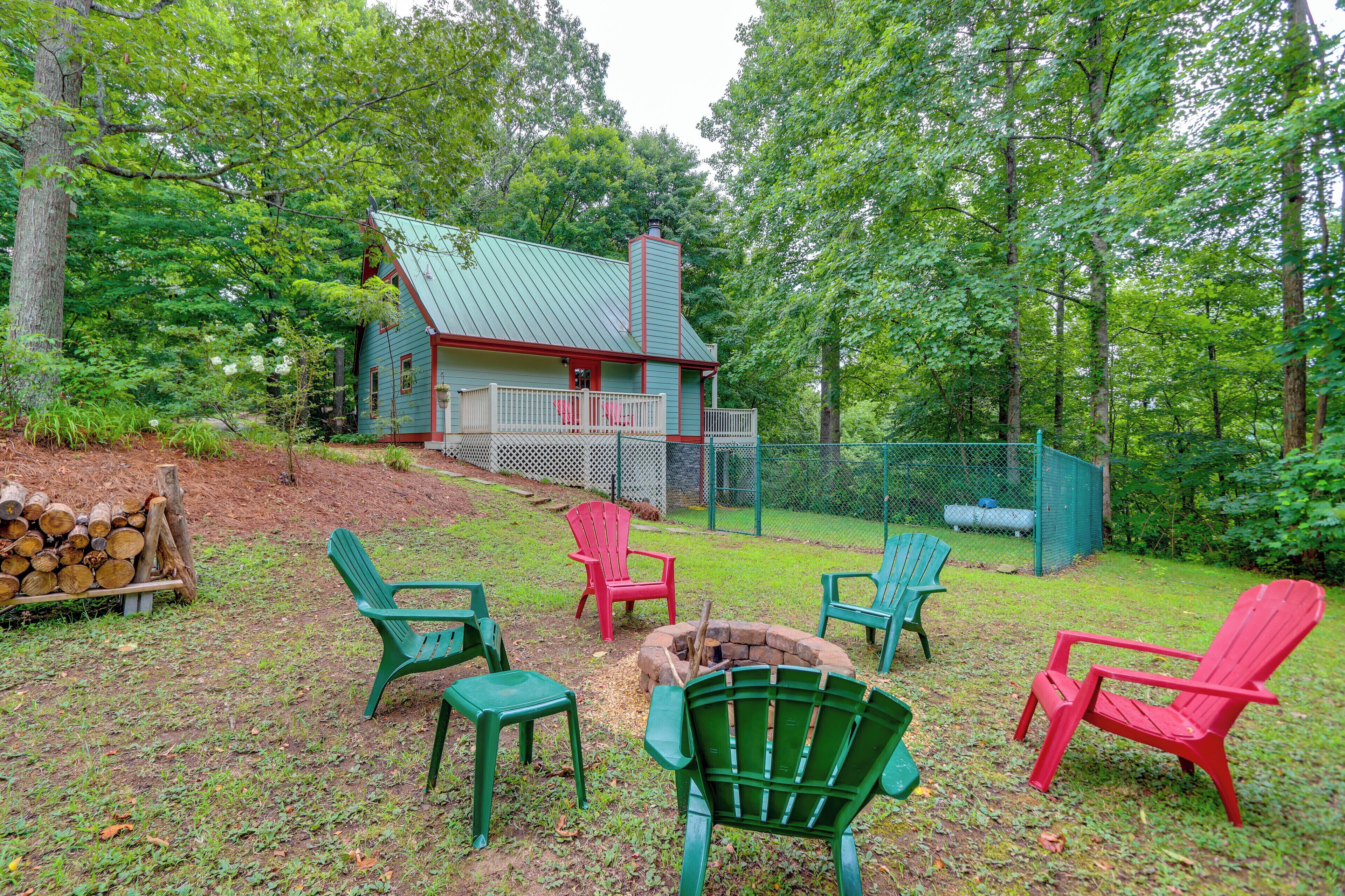 A cabin with a green roof and red trim, surrounded by a forest. There are four Adirondack chairs, two red and two green, arranged around a fire pit.