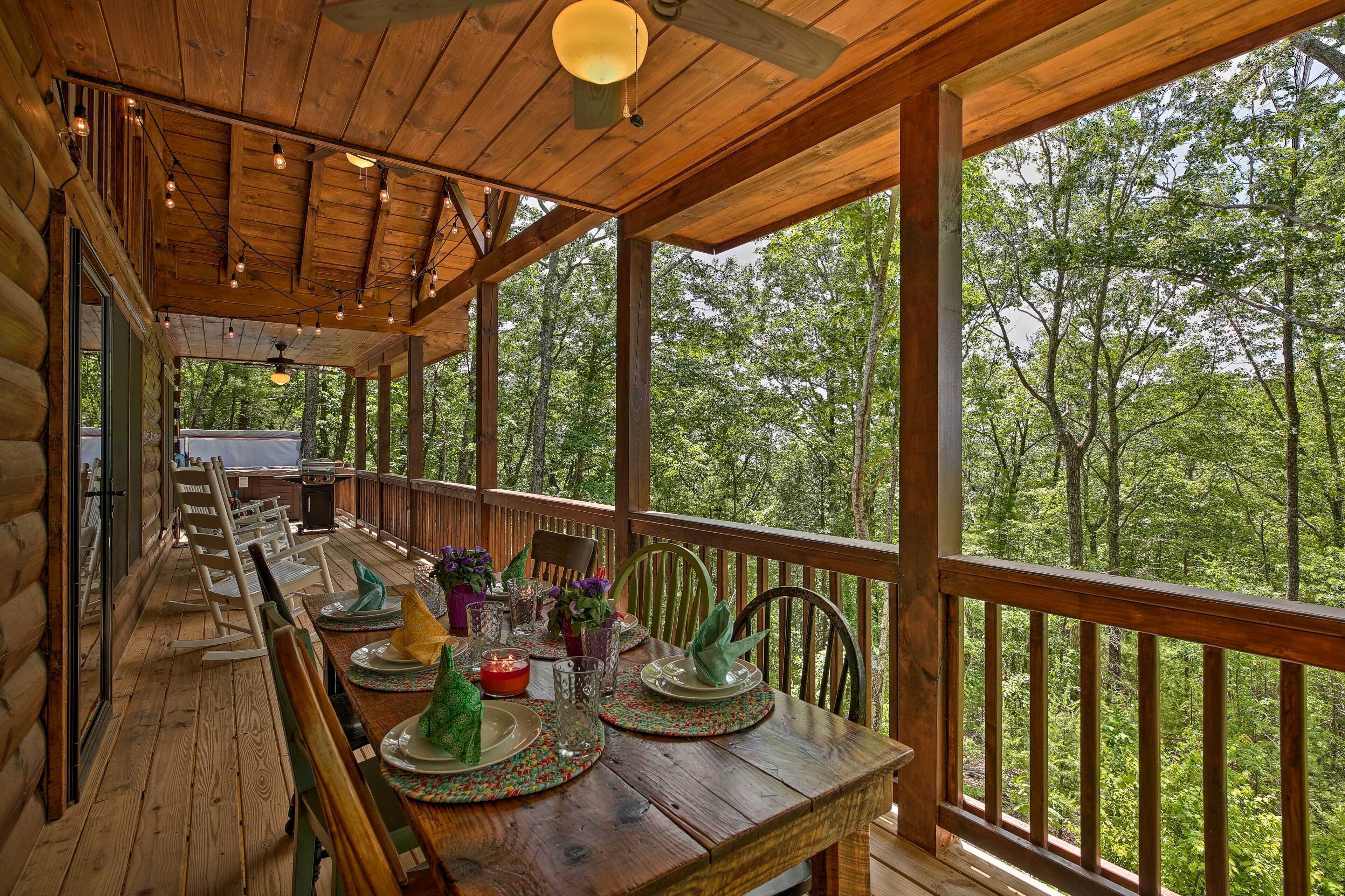 A wooden deck with a long table set for dining, surrounded by chairs, and overlooking a forested area.