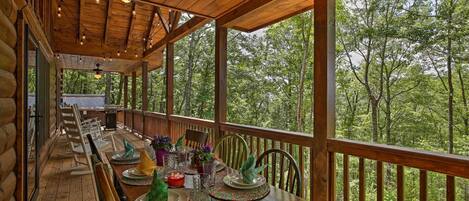 A wooden deck with a long table set for dining, surrounded by chairs, and overlooking a forested area.