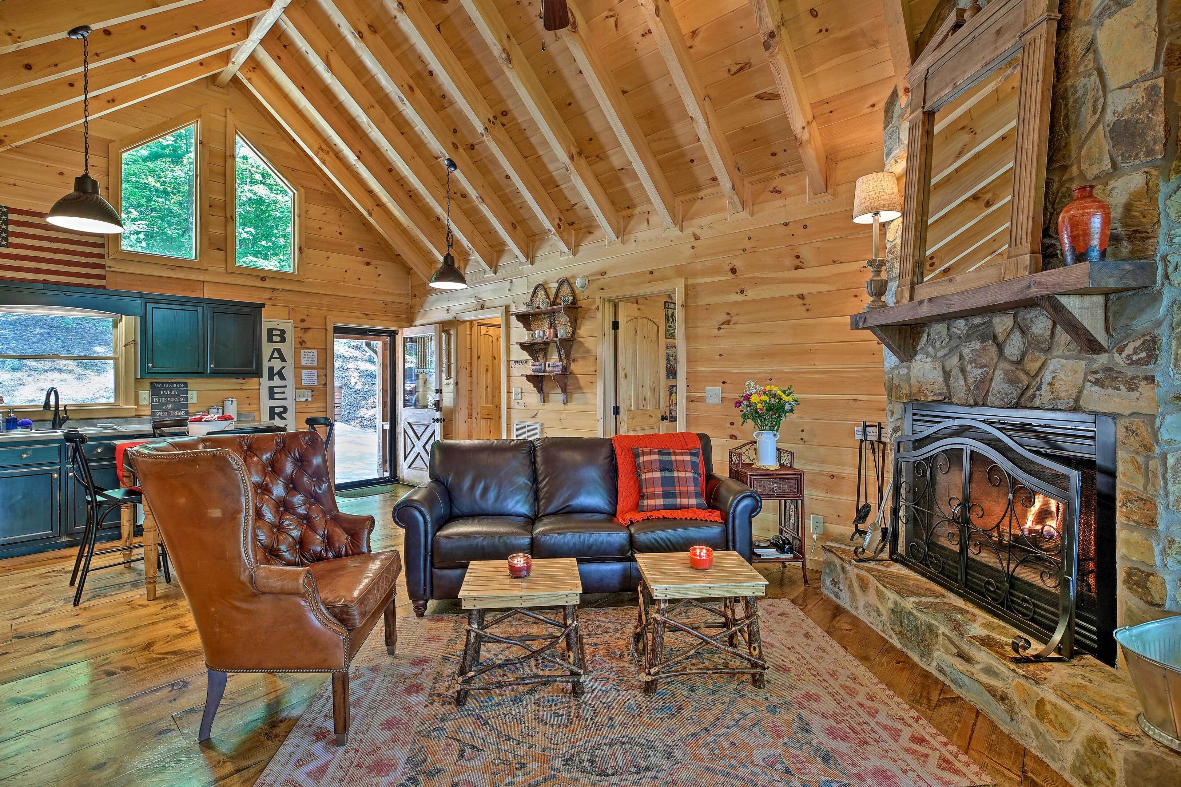 A cozy cabin interior with a leather sofa, a stone fireplace, and a kitchen area visible through the windows.