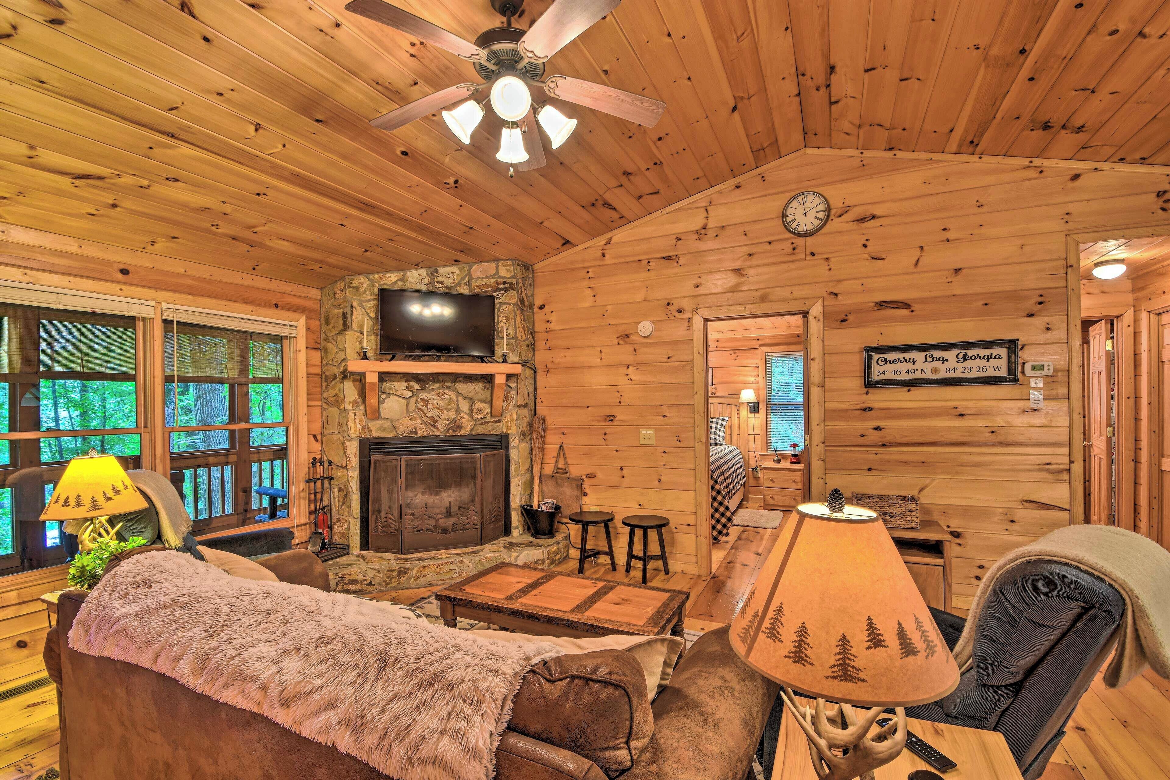 A cozy cabin interior with a stone fireplace, wooden walls, a ceiling fan, and a television mounted above it.