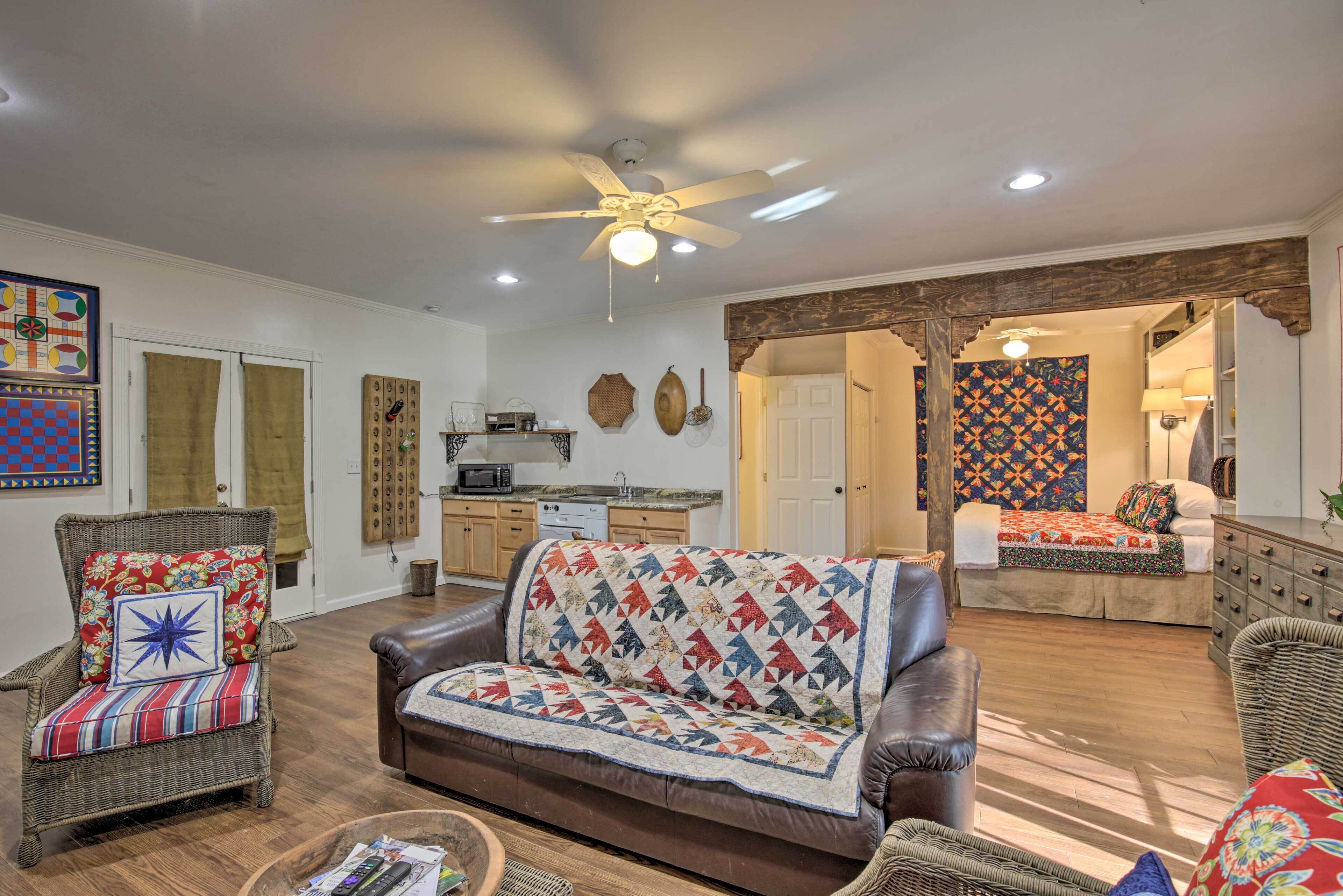 A living room with a brown leather sofa, a wicker armchair, and a quilted throw. There is a ceiling fan, a kitchen area, and a door leading to another room.