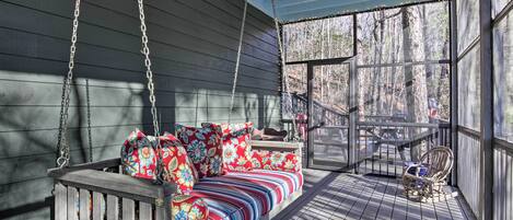 A covered porch with a wooden swing seat and cushions, a wicker chair, and a view of the surrounding forest.