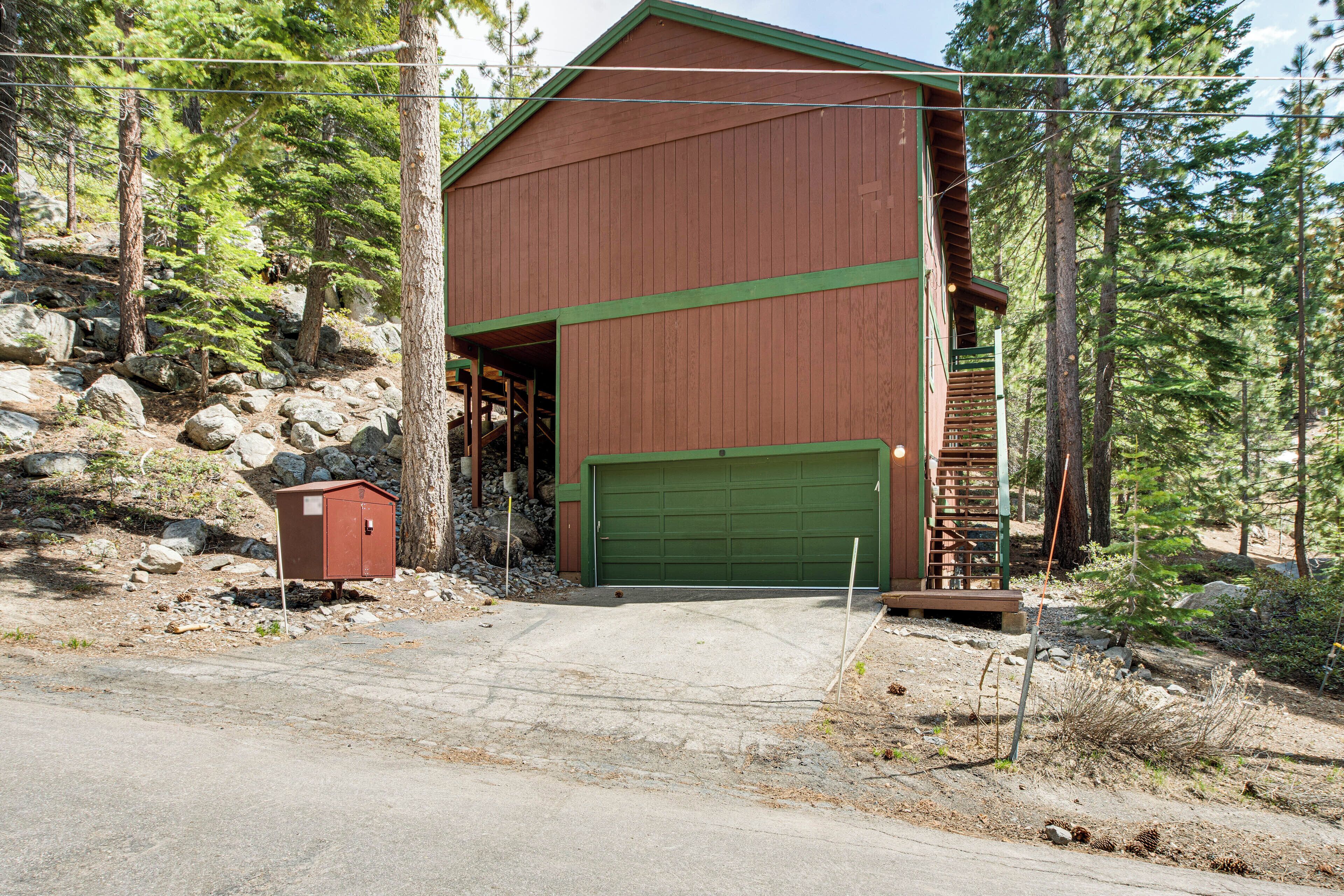 A two-story wooden building with a green garage door, surrounded by trees and rocks.