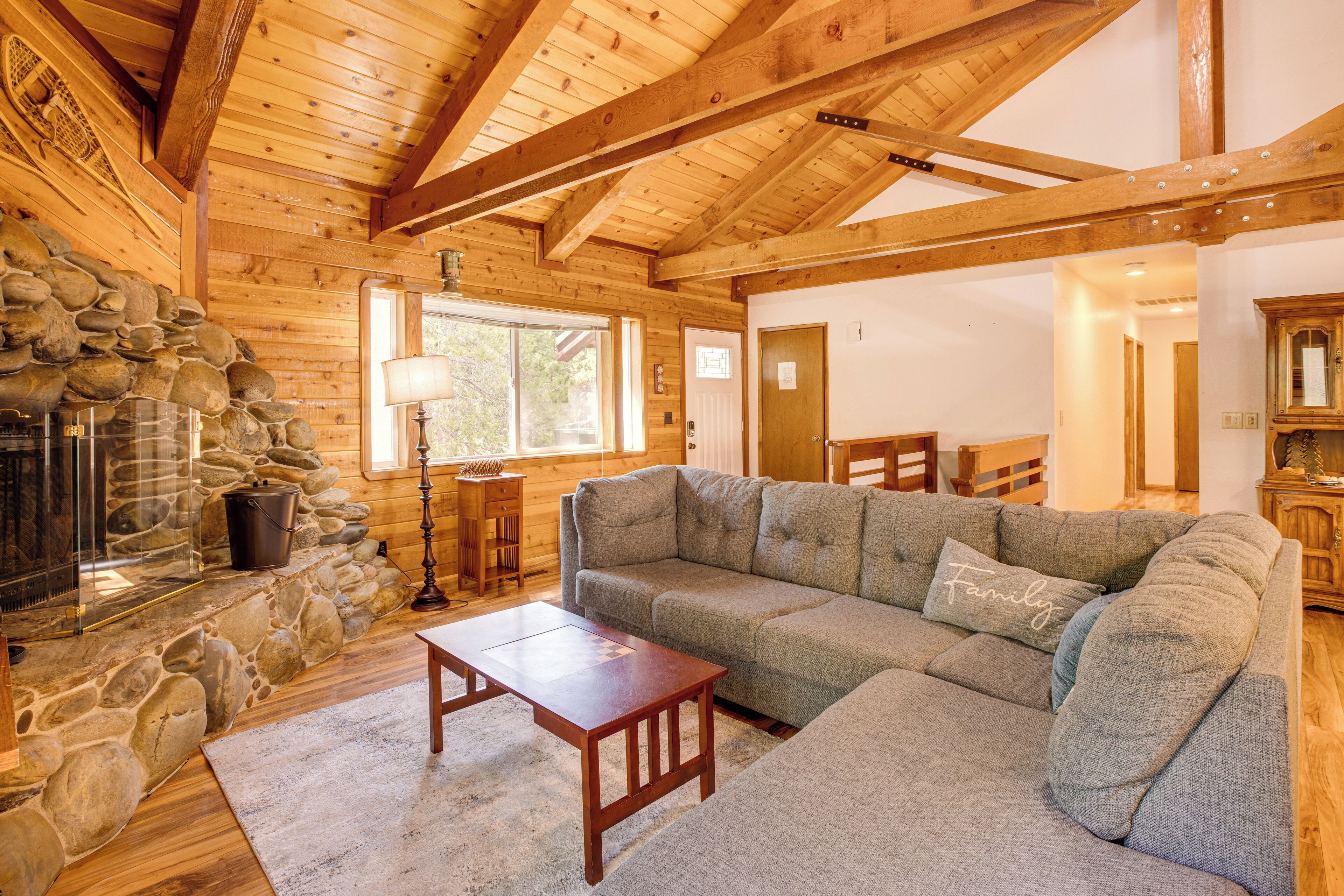 A cozy log cabin interior with a stone fireplace, wooden beams, a gray sofa, and a small wooden table.