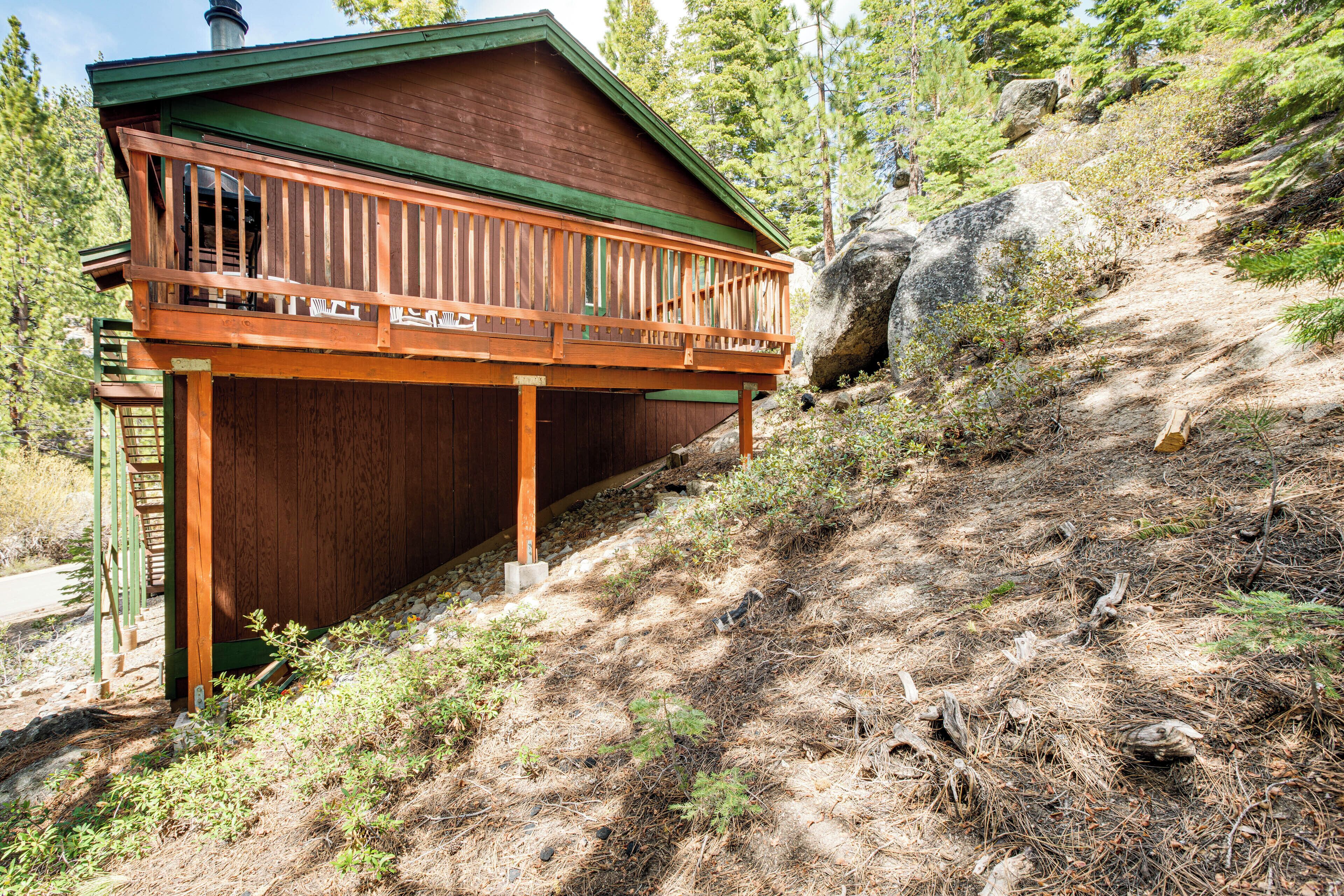A wooden cabin with a balcony, surrounded by trees and a rocky hillside.