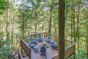 Um deck de madeira com cadeiras Adirondack e uma lareira, tudo cercado por uma floresta.