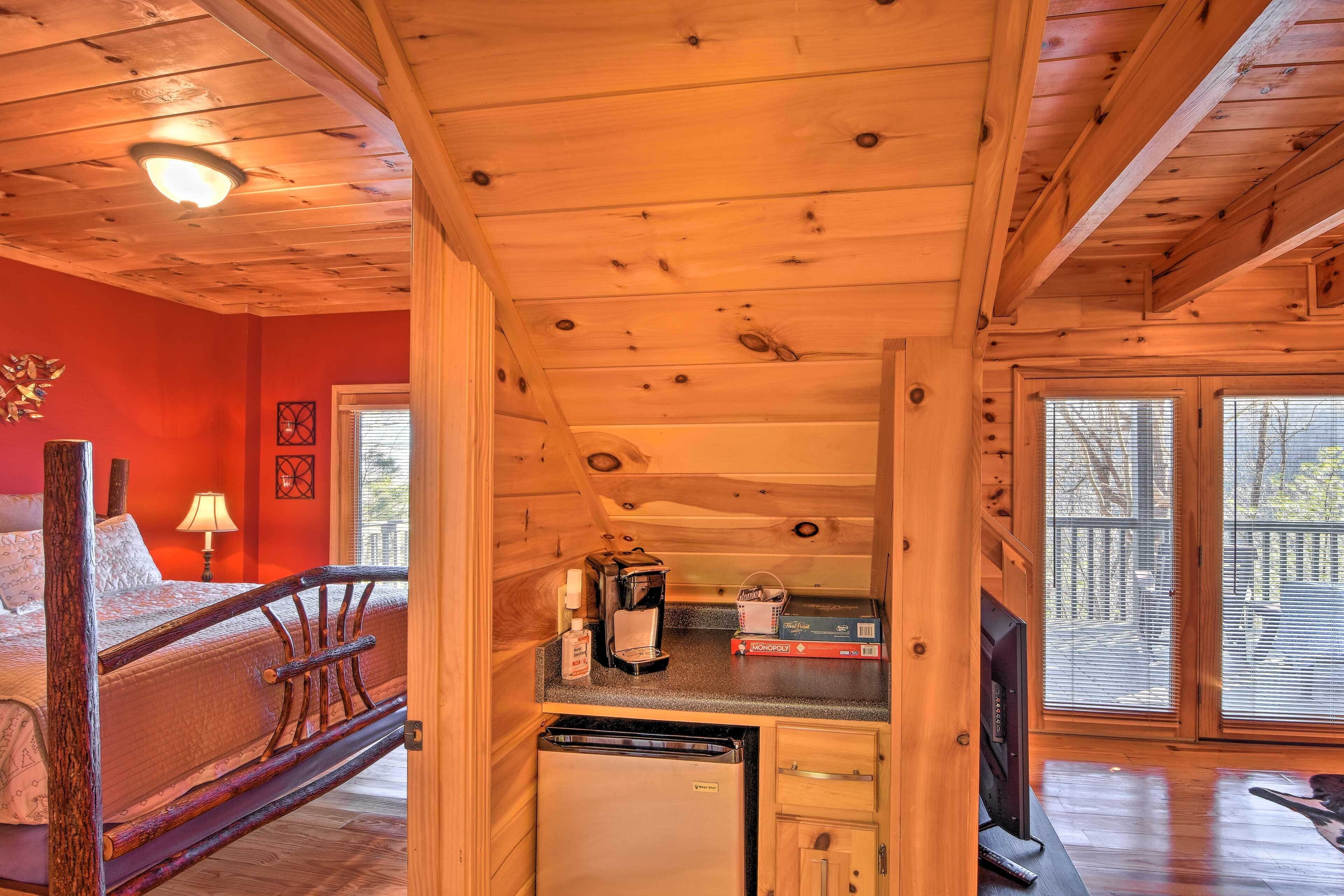 A cozy cabin interior with a wooden ceiling, a bed with a headboard, a kitchen area with a refrigerator, and a large sliding glass door leading to an outdoor deck.