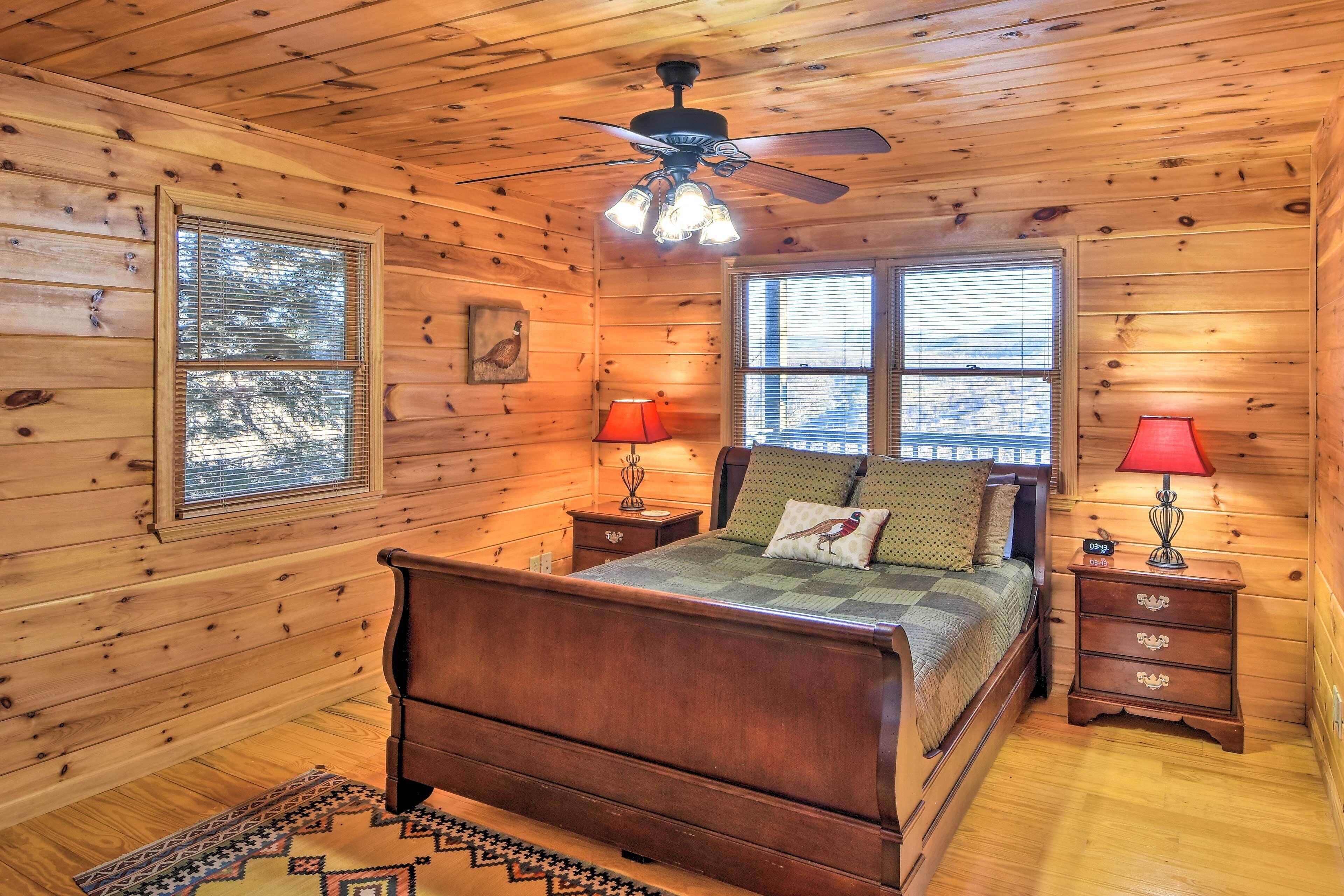 A wooden cabin bedroom with a bed, two bedside tables, a ceiling fan, and two windows.