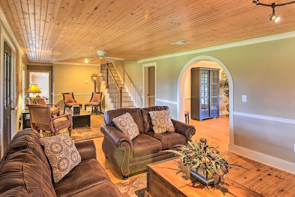 A spacious living room with a brown leather sofa set, a coffee table with a floral arrangement, and a staircase leading to another level.