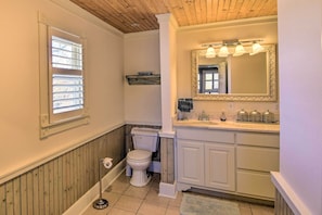 A bathroom with a wooden ceiling, a toilet, a sink with a mirror, and a window with blinds. - Serving Southern Charm at This Clarkesville House! (Clarkesville)