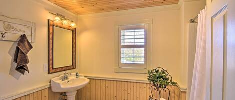 A bathroom with a wooden ceiling, a white sink, a mirror, a window with blinds, and a towel rack.