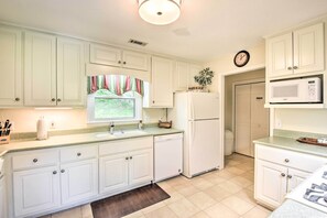A kitchen with white cabinets, a microwave, a refrigerator, a clock, and a window with curtains.