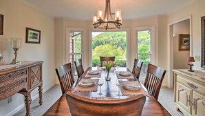 A dining room with a large wooden table set for six, surrounded by chairs, a chandelier, and a view of a lush garden through large windows. - Sky Valley Retreat w/ Resort Amenities & Views! (Dillard)
