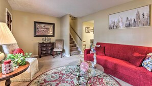 Ein Wohnzimmer mit einem roten Sofa, einem Glastisch, einem gemusterten Teppich, einer Lampe und einer Treppe, die in ein weiteres Zimmer führt.