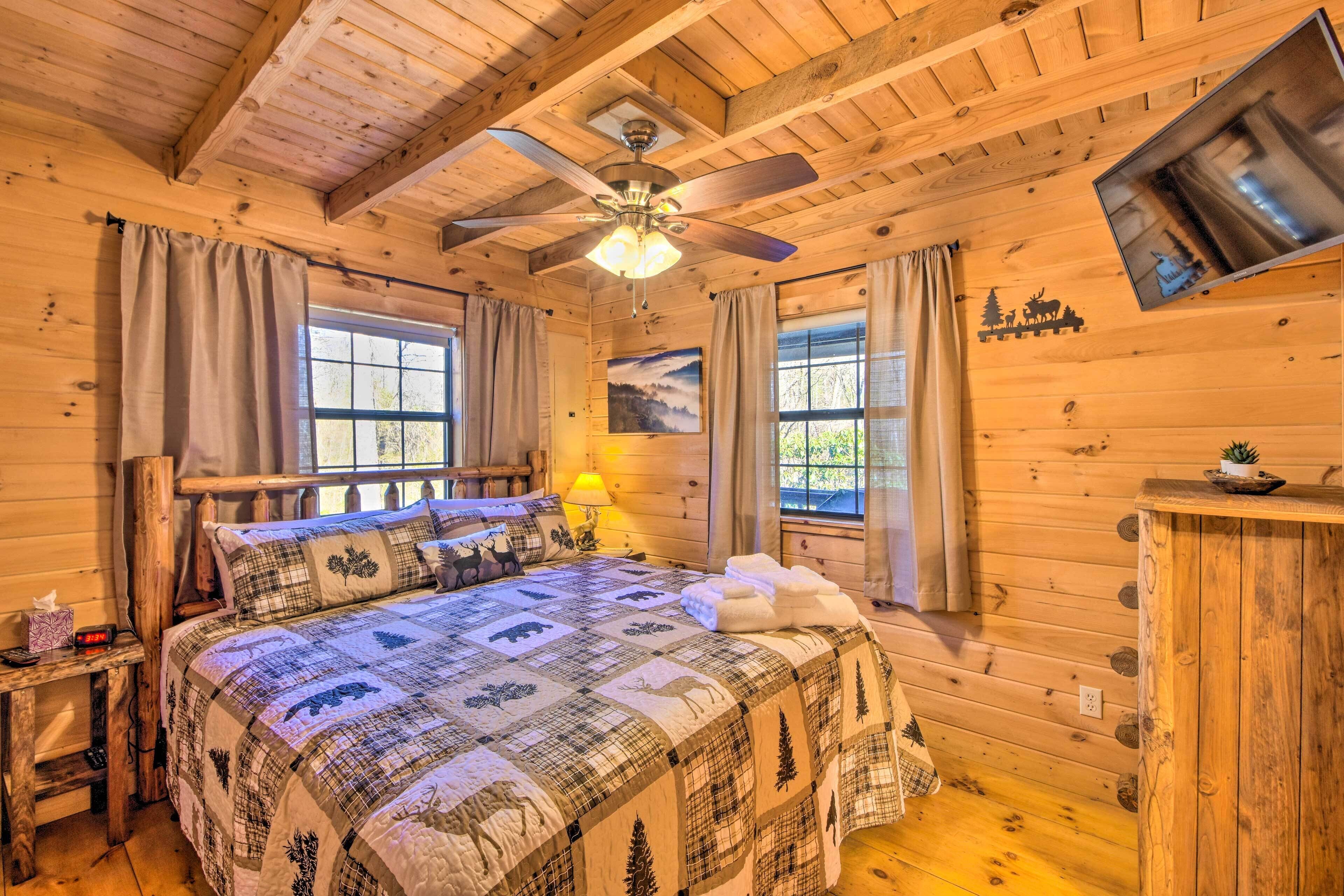 A cozy log cabin bedroom with a large bed, a ceiling fan, and a TV mounted on the wall.
