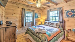 Une chambre confortable dans un chalet, avec un intérieur en bois, un lit recouvert d’une courtepointe, un ventilateur de plafond et deux fenêtres avec des rideaux.