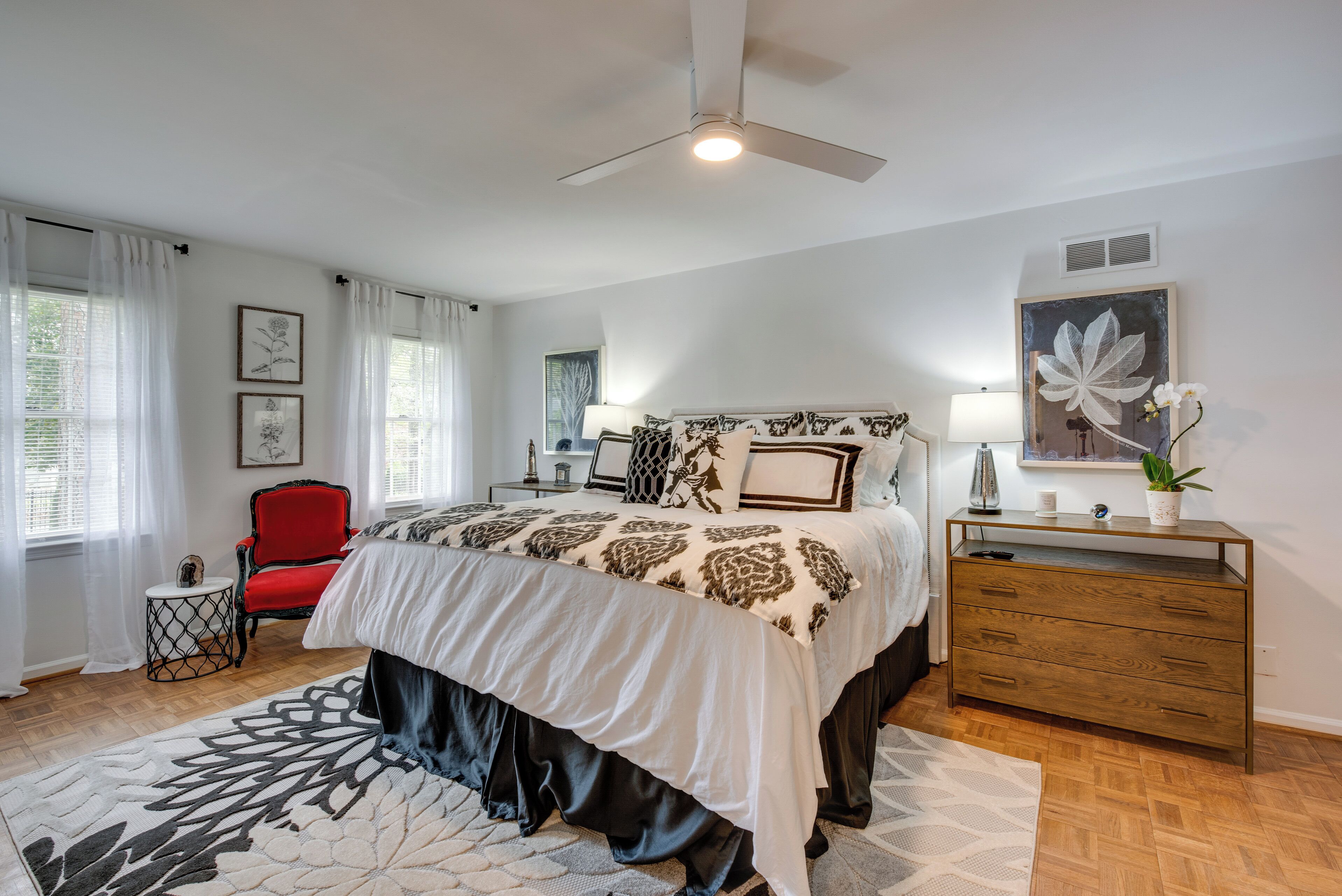 A bedroom with a bed, a red chair, a wooden dresser, and a ceiling fan.