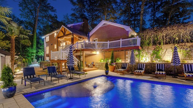 A swimming pool with lounge chairs and umbrellas, surrounded by a house with a balcony and a stone wall. - Luxury Retreat in Smyrna: Pool, Pickleball & More! (Smyrna)