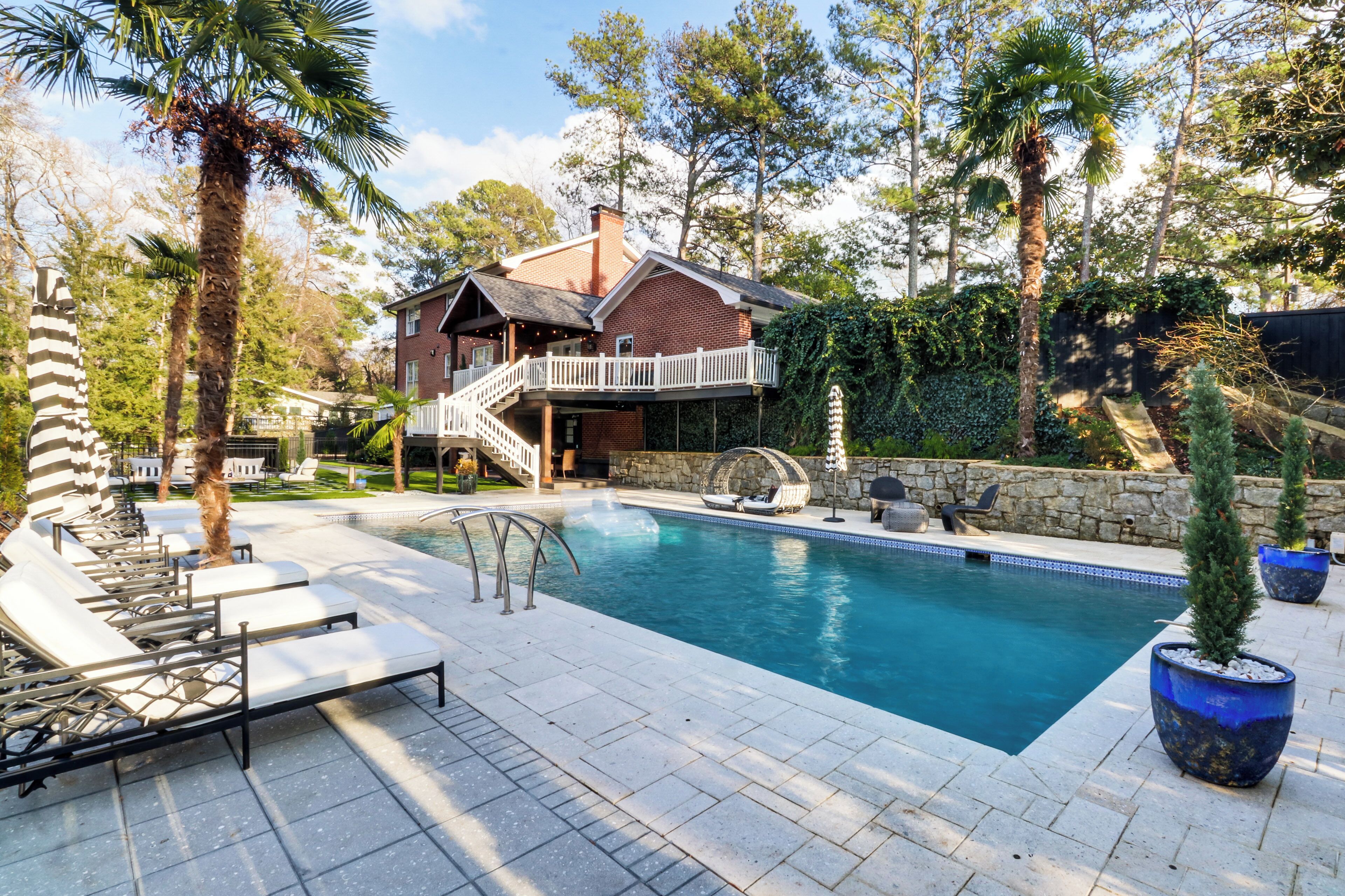 Una piscina con sillas de descanso, una casa de ladrillo con balcón y abundante vegetación.