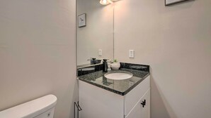 A bathroom with a white toilet, a granite countertop sink, a mirror with lights, and a framed picture on the wall.