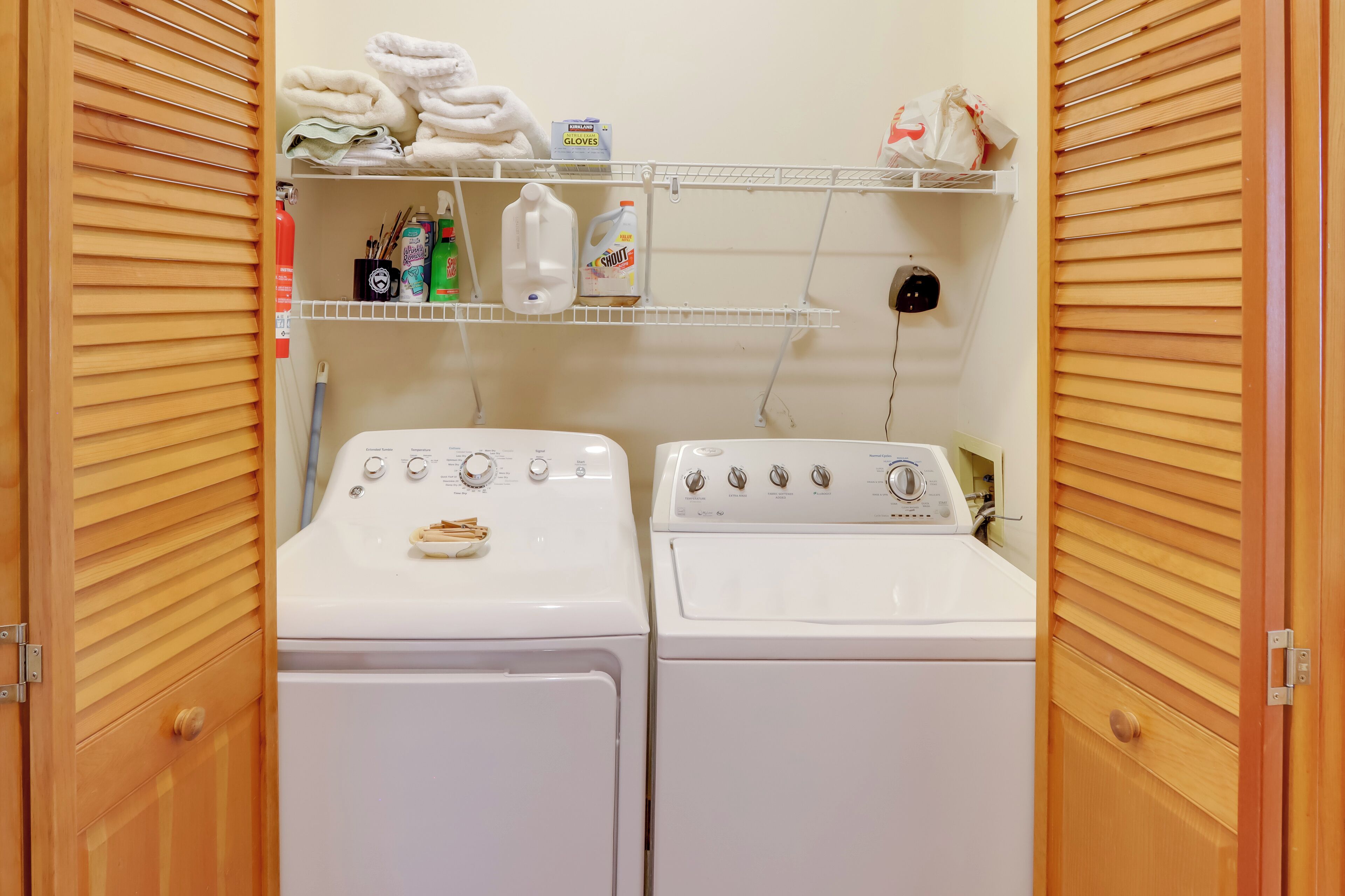 A laundry room with two white front-loading washing machines, shelves with towels, cleaning supplies, and a fire extinguisher.