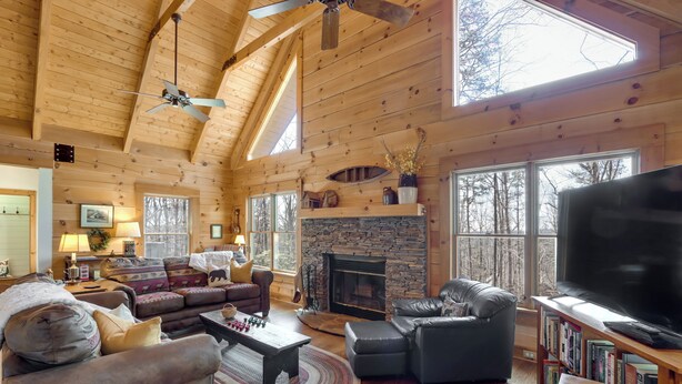 L’intérieur chaleureux d’un chalet en bois comprend une cheminée, des canapés en cuir, un téléviseur à écran plat et une grande fenêtre donnant sur la forêt.
