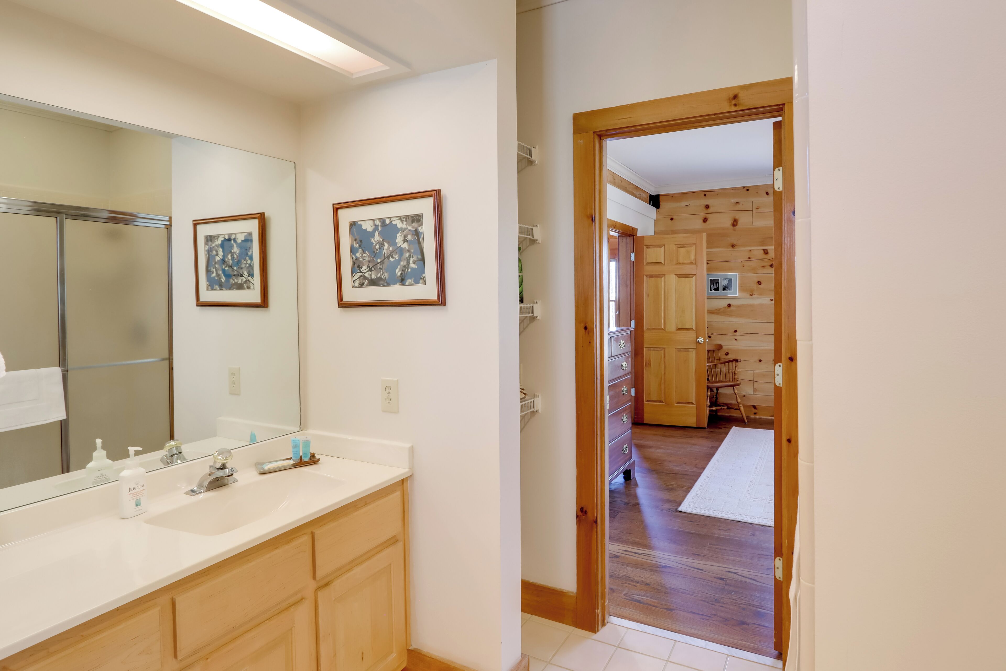 A bathroom with a large mirror, two framed artworks, and a white sink with toiletries.
