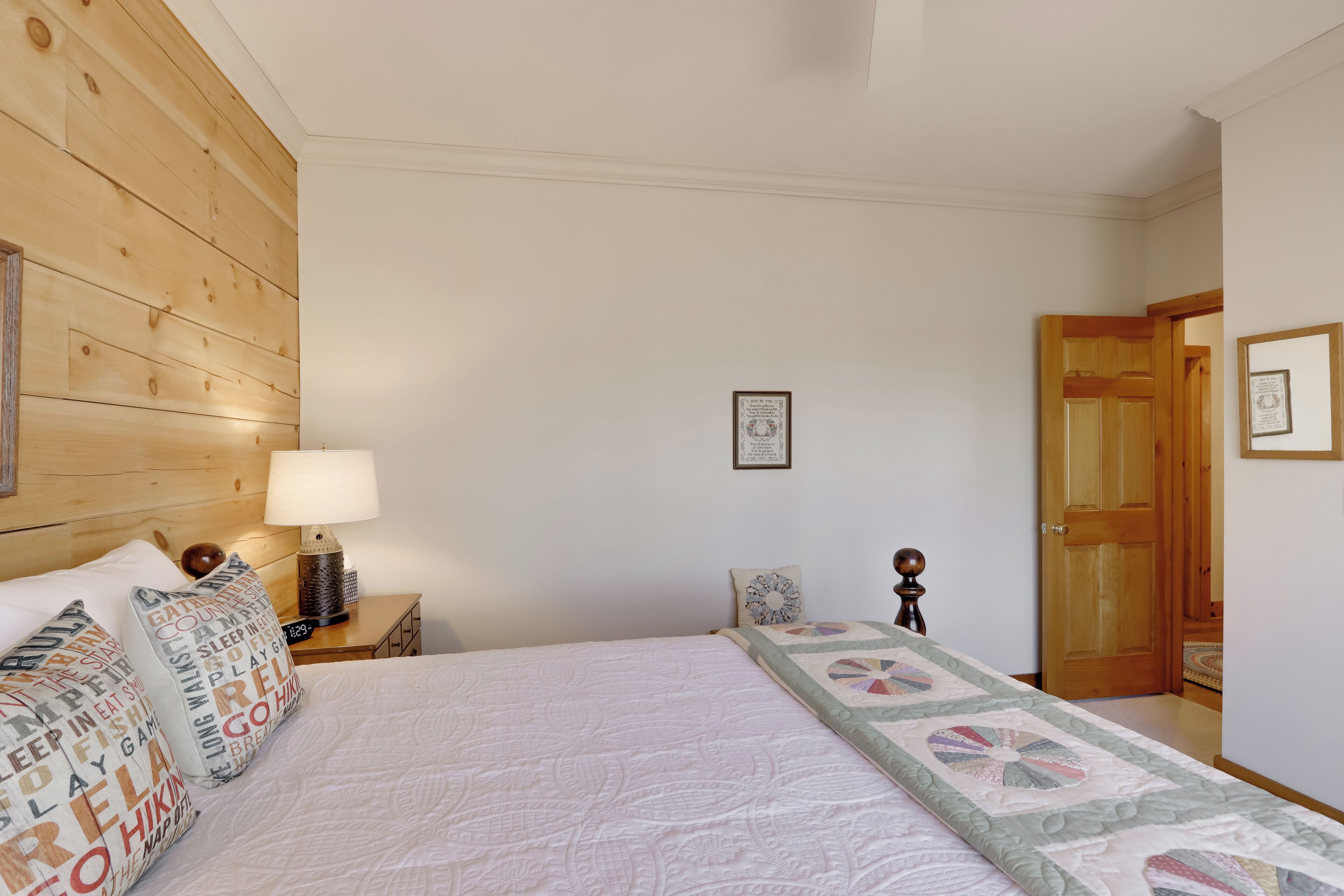 A bedroom with a wooden headboard, a bed with patterned pillows, a bedside table with a lamp, and a door leading to another room.