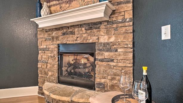 Une cheminée en pierre avec une étagère blanche au-dessus du foyer, un feu qui crépite à l’intérieur, et une bouteille de vin et un verre posés sur le côté droit.