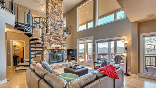 A spacious living room with a stone fireplace, a large leather sofa, and a staircase leading to an upper level. - Sky Valley Home w/ Mtn View, 5 Mi to Rabun Bald! (Dillard)