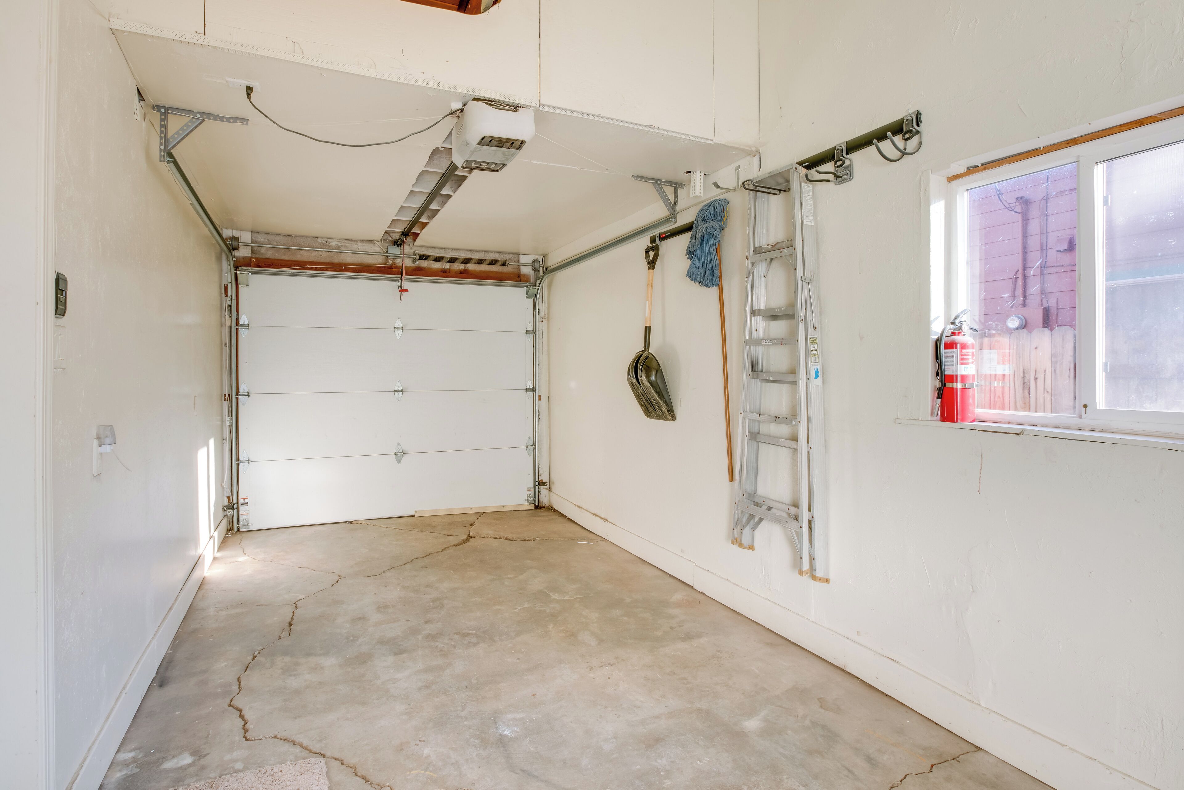 A garage with a concrete floor, a white door, and a window with a view of a red fence.